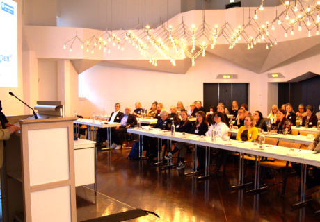 Sitzungssaal mit Teilnehmenden der Tagung und am Podium steht eine Frau und begrüßt